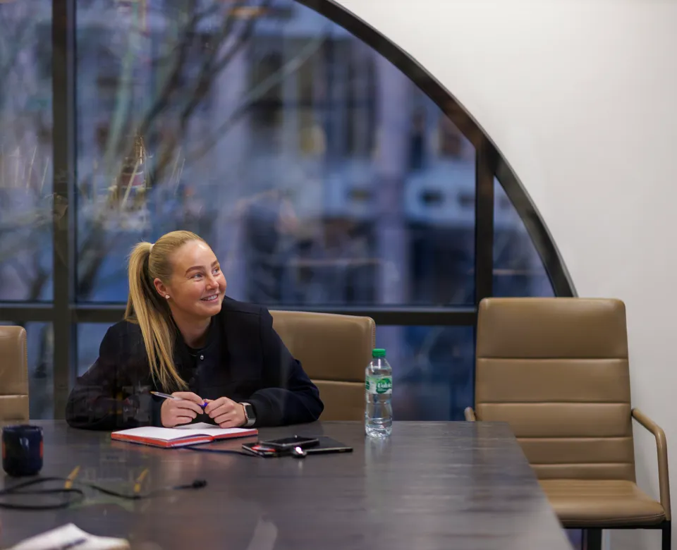 Smiling woman in a black blazer sitting at a conference table with a notepad.