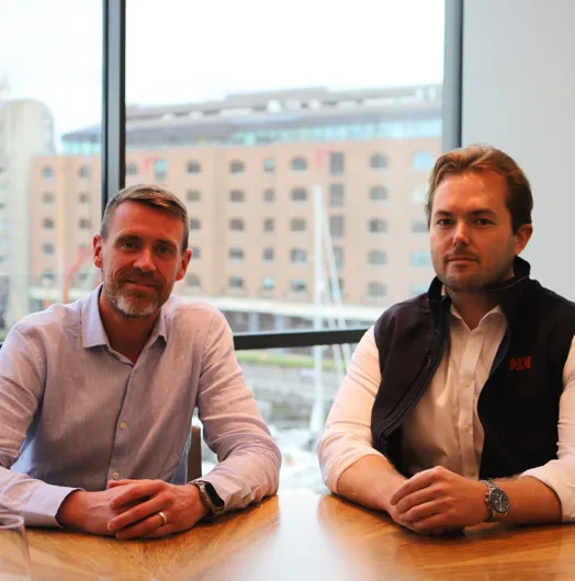 Two men seated at a wooden table in front of a large window overlooking a marina.