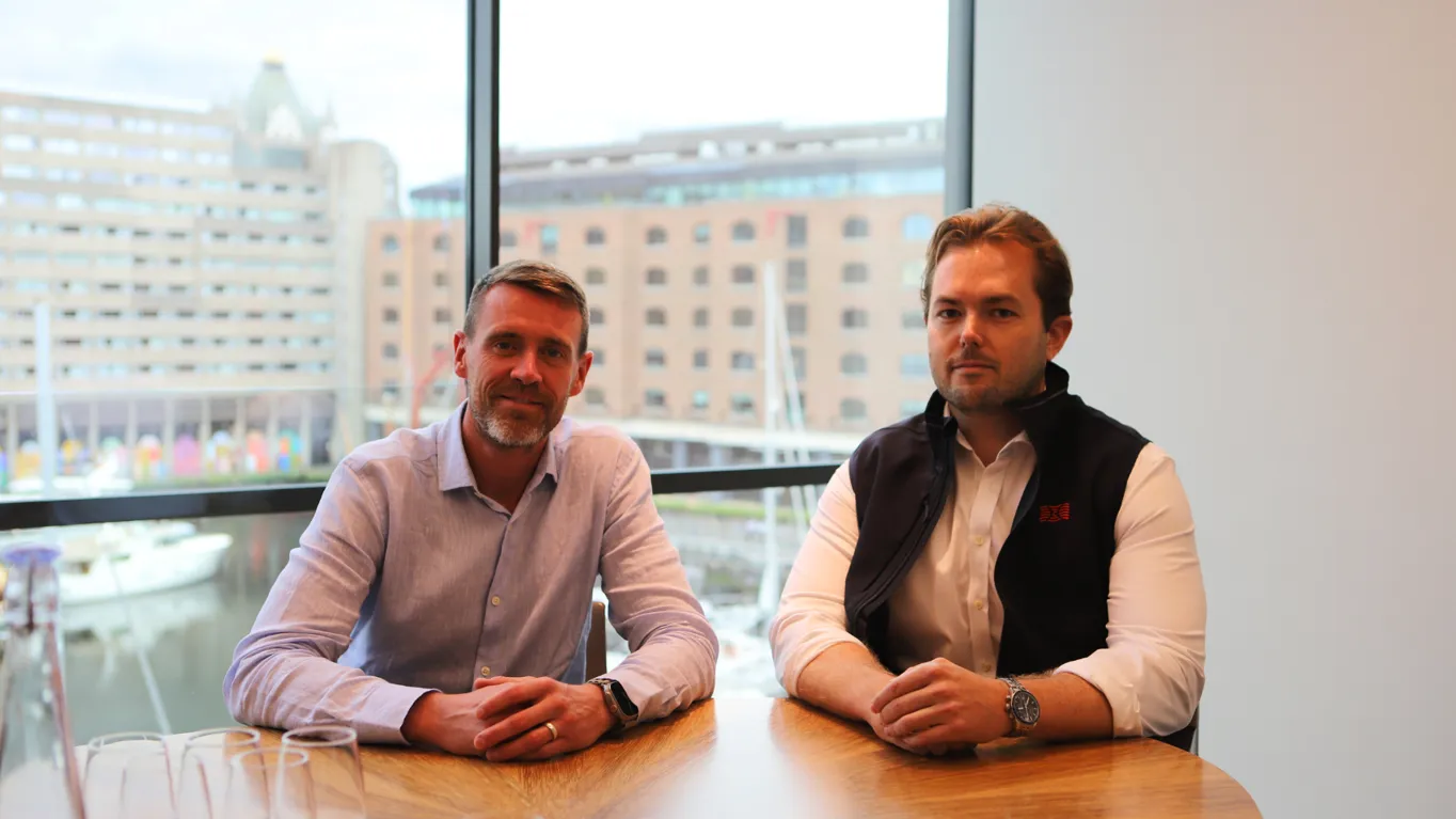 Two men seated at a wooden table in front of a large window overlooking a marina.