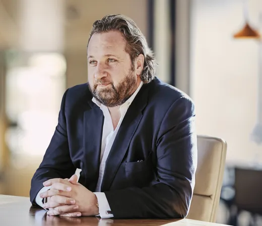 Man with a beard in a dark suit seated at a table, deep in thought, in an office setting.