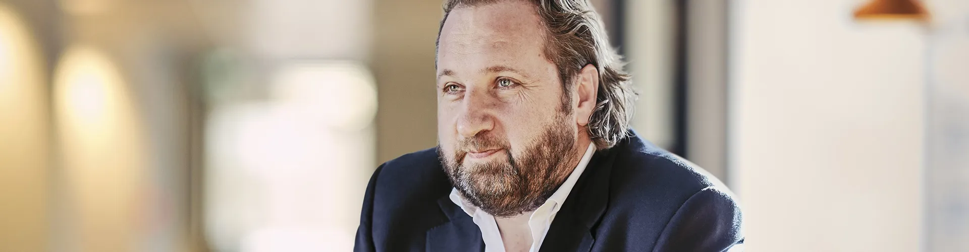 Man with a beard in a dark suit seated at a table, deep in thought, in an office setting.