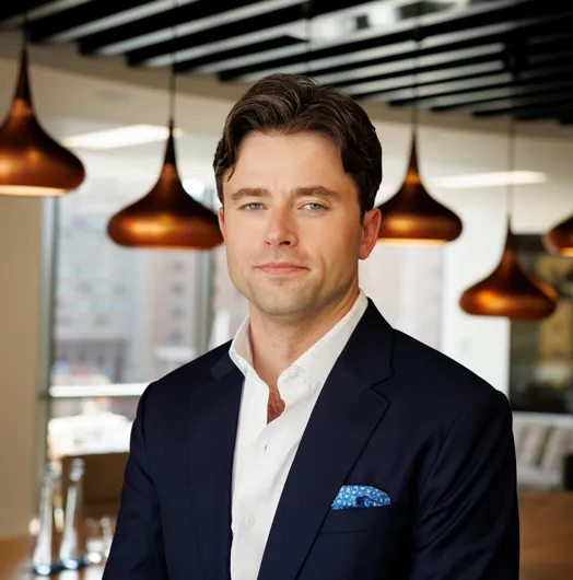 Man in a navy suit jacket with a pocket square standing indoors with an urban background.
