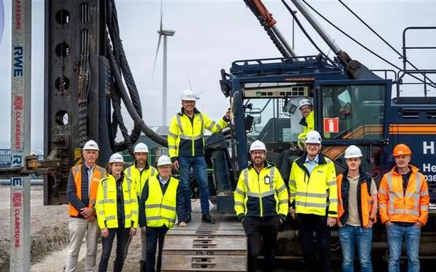 Eleven construction workers in safety gear posing by heavy machinery with a wind turbine.