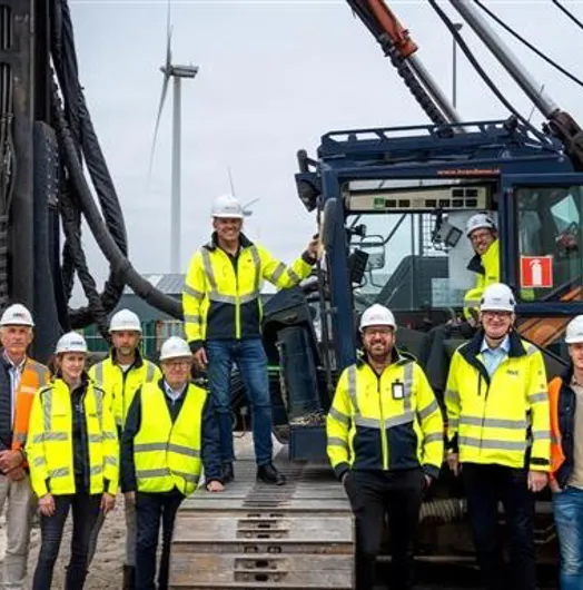 Eleven construction workers in safety gear posing by heavy machinery with a wind turbine.