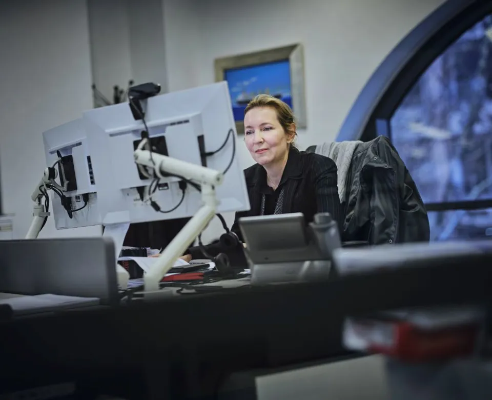 Woman in a dark jacket sitting at a desk, looking at a computer monitor with a snowy view outside.