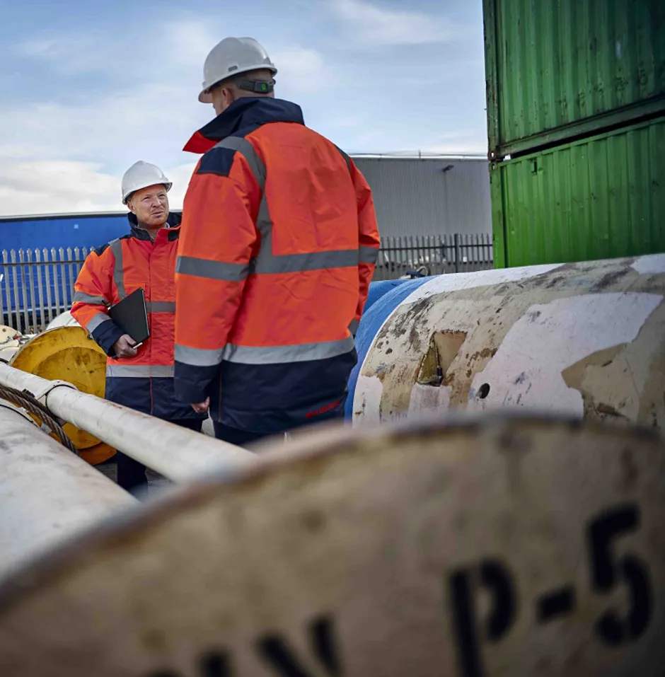 Two men in safety helmets and orange jackets talking among large pipes and equipment.
