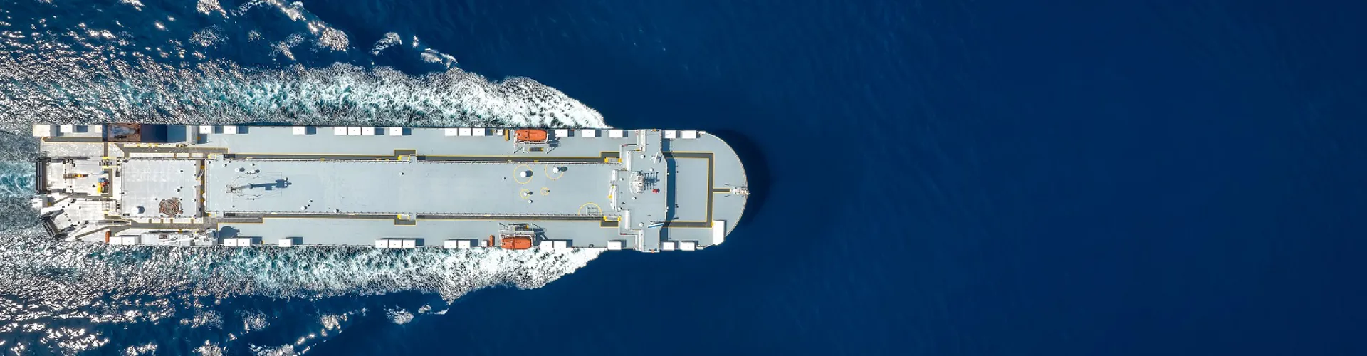 Aerial view of a large cargo ship moving through deep blue water, leaving a white wake.