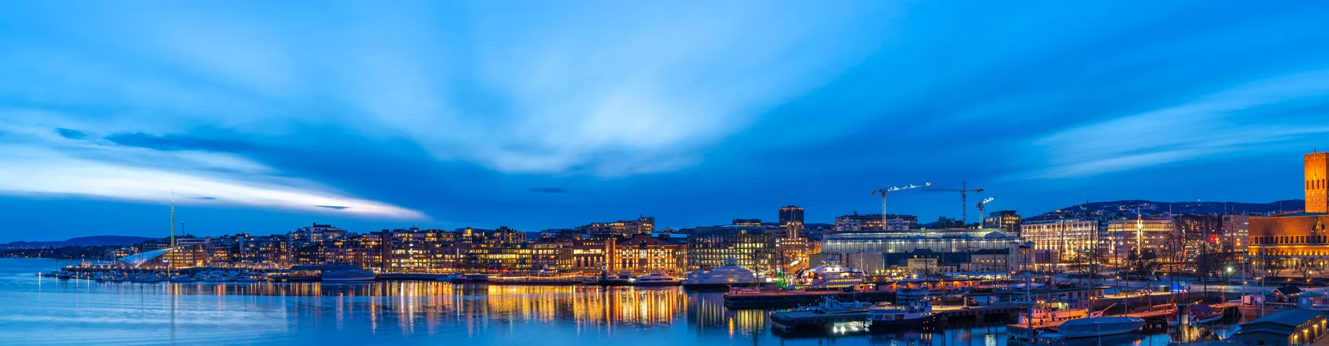 Panoramic view of a harbor city at dusk with modern waterfront buildings, reflections, and boats.
