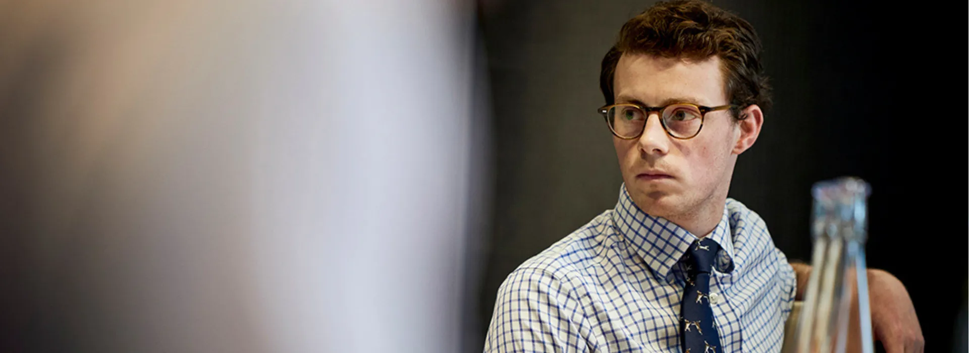 Man in checked shirt and tie, seated, looking serious.