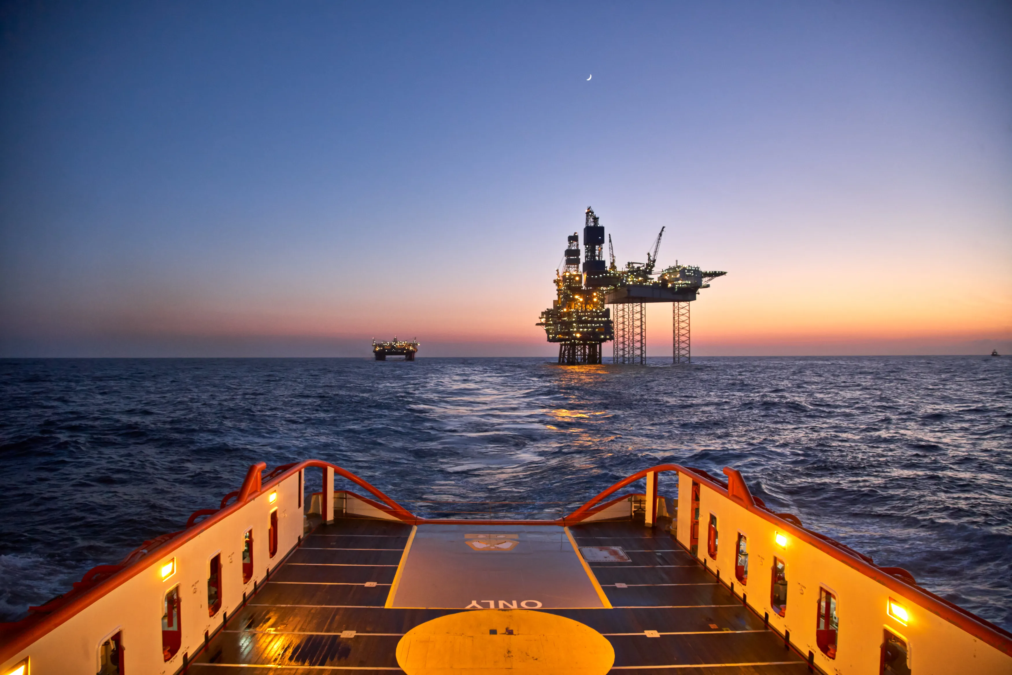View from a ship deck of two illuminated oil rigs in the ocean at sunset with a crescent moon.