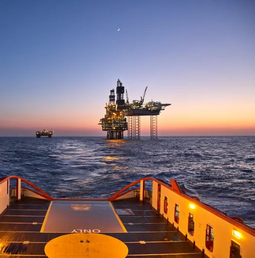 View from a ship deck of two illuminated oil rigs in the ocean at sunset with a crescent moon.