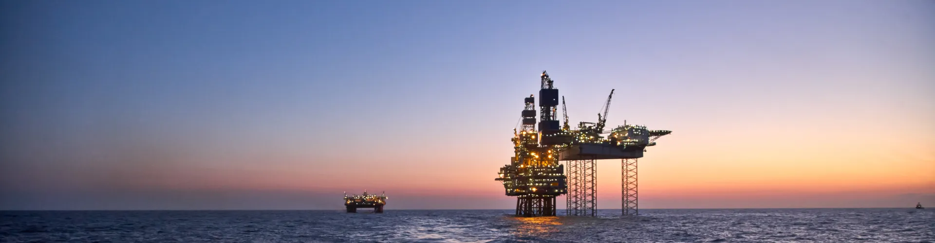 View from a ship deck of two illuminated oil rigs in the ocean at sunset with a crescent moon.