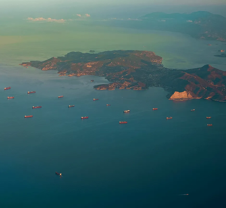 Aerial view of a coastal area with cargo ships, a mountainous island, and a tranquil blue sea.