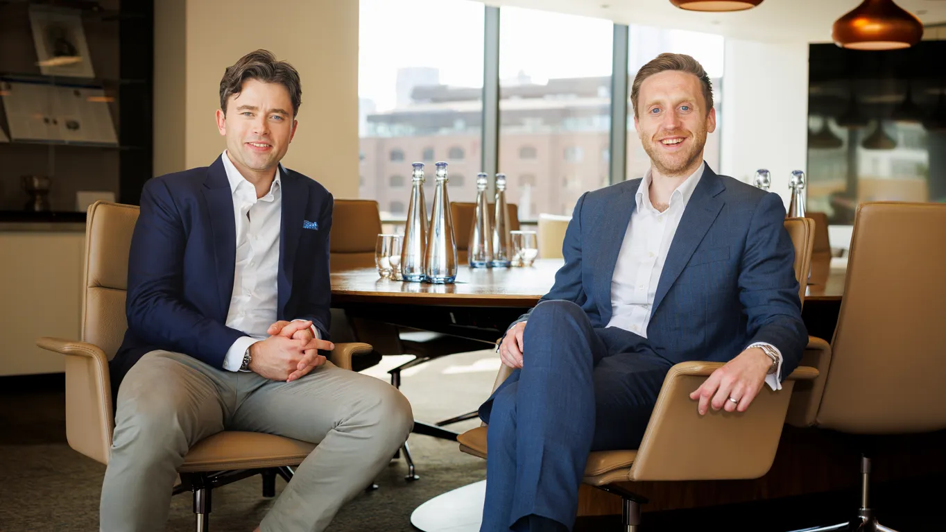 Two men in suits sitting in an office, smiling.